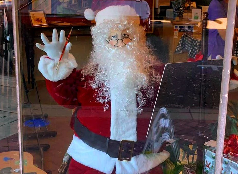 Santa sits at an easel with paintbrush in hand and palette of paint nearby